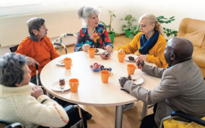 Elderly people sitting around a table eating cake and talking. Some with mobility aids.