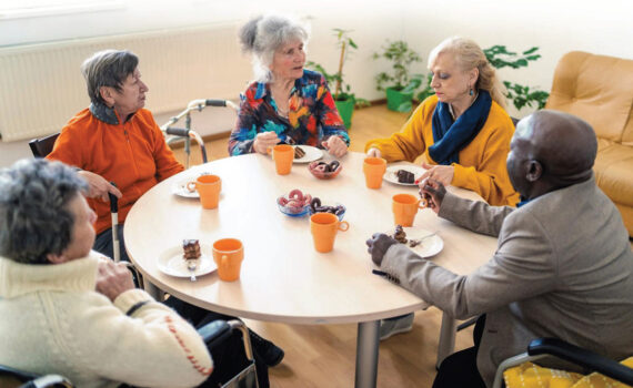 Elderly people sitting around a table eating cake and talking. Some with mobility aids.