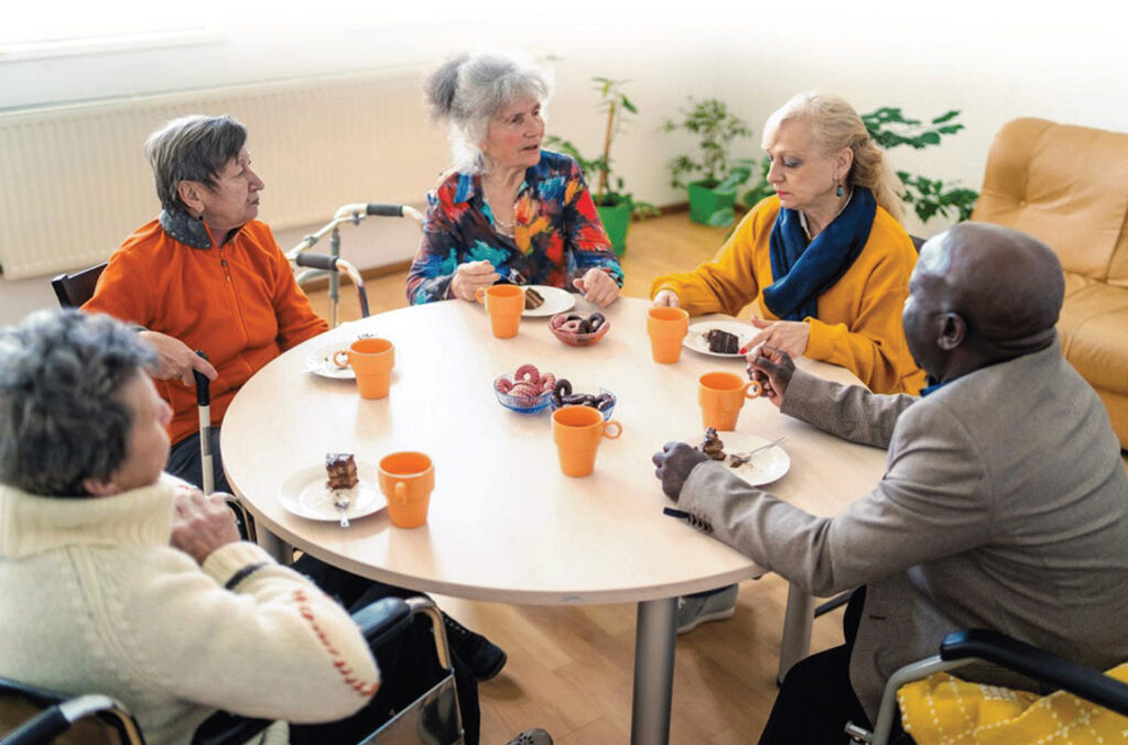 Elderly people sitting around a table eating cake and talking. Some with mobility aids.